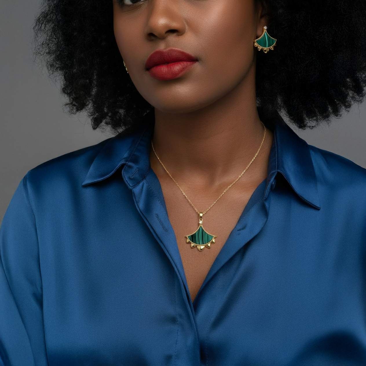 A woman wearing a blue satin blouse models matching gold and malachite dome earrings and a pendant necklace.